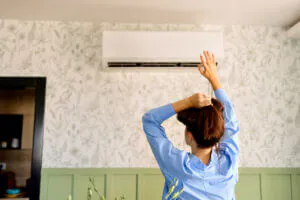 AC emergency signs: A woman stands indoors, reaching up toward an air conditioner and checking if it’s working properly.
