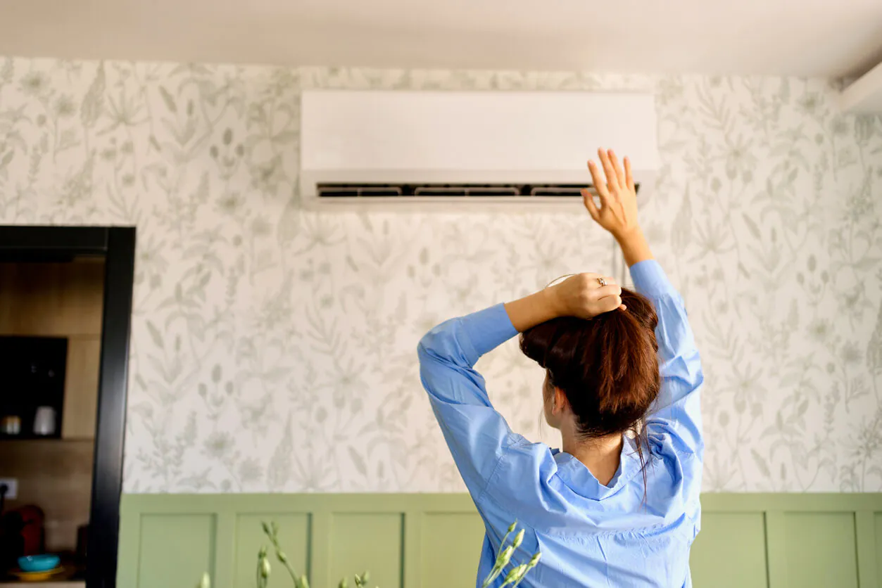 AC emergency signs: A woman stands indoors, reaching up toward an air conditioner and checking if it’s working properly.